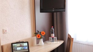 Habitación de hotel con un escritorio de madera, una silla, un televisor de pantalla plana montado en la pared, una tableta sobre el escritorio, un jarrón con flores y una botella de agua.