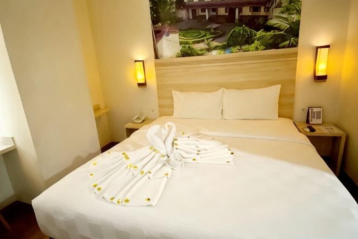 A hotel room with a bed, two bedside lamps, a telephone, and a painting of a tropical landscape.