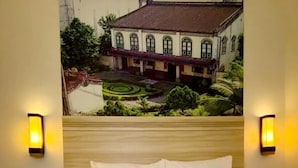 A hotel room with a bed featuring a towel arrangement in the shape of a swan, two wall-mounted lamps, and a mural of a building and garden on the wall. - Cordela Inn Millennium Medan (Dwi Kora)