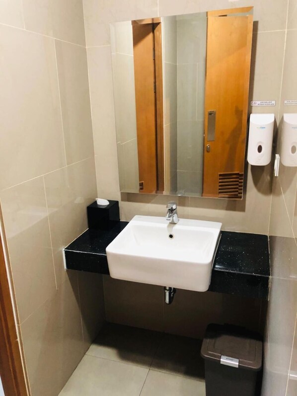 A bathroom with a white sink, a mirror, a trash bin, and a soap dispenser. - Cordela Inn Millennium Medan (Dwi Kora)