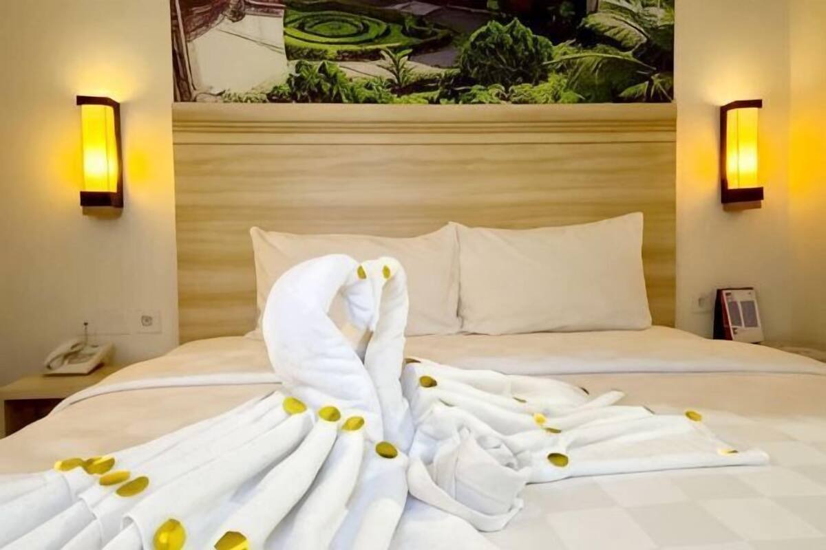 Hotellihuone, jossa on sänky, kaksi seinälle asennettua lamppua, sängyn yläpuolella oleva taulu ja yöpöytä, jossa on puhelin.