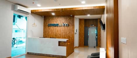 A hotel reception area with a wooden reception desk, a seating area with a lamp, and a view of the street outside.