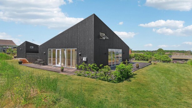 Un edificio moderno nero con ampie porte in vetro e una piccola finestra, immerso nel verde e con un'area pavimentata in legno.
