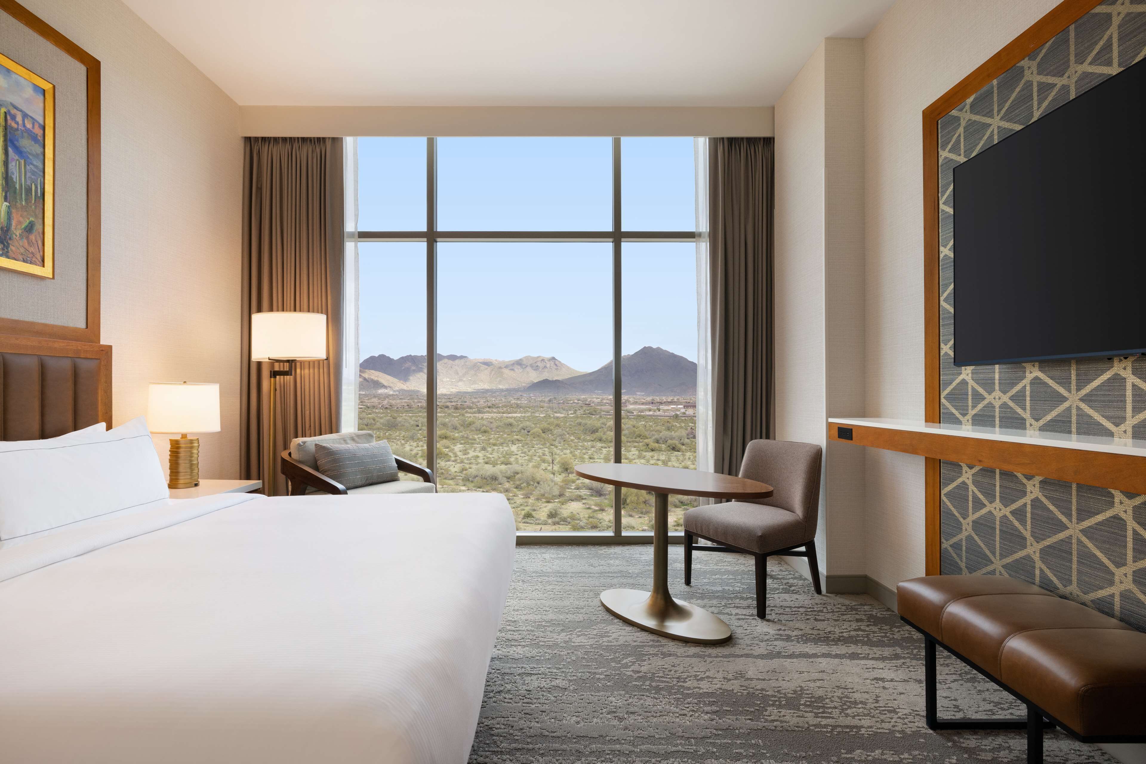 Habitación de hotel con una cama grande, un escritorio, una silla, un banco y vistas a las montañas a través de amplias ventanas.