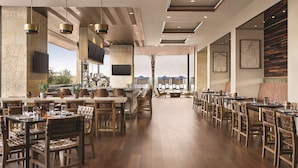 A spacious restaurant with wooden flooring, high tables, and chairs, featuring a bar area and large windows overlooking a scenic view.