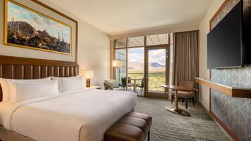 A hotel room with a large bed, a desk with a chair, a TV mounted on the wall, and a window with a mountain view.