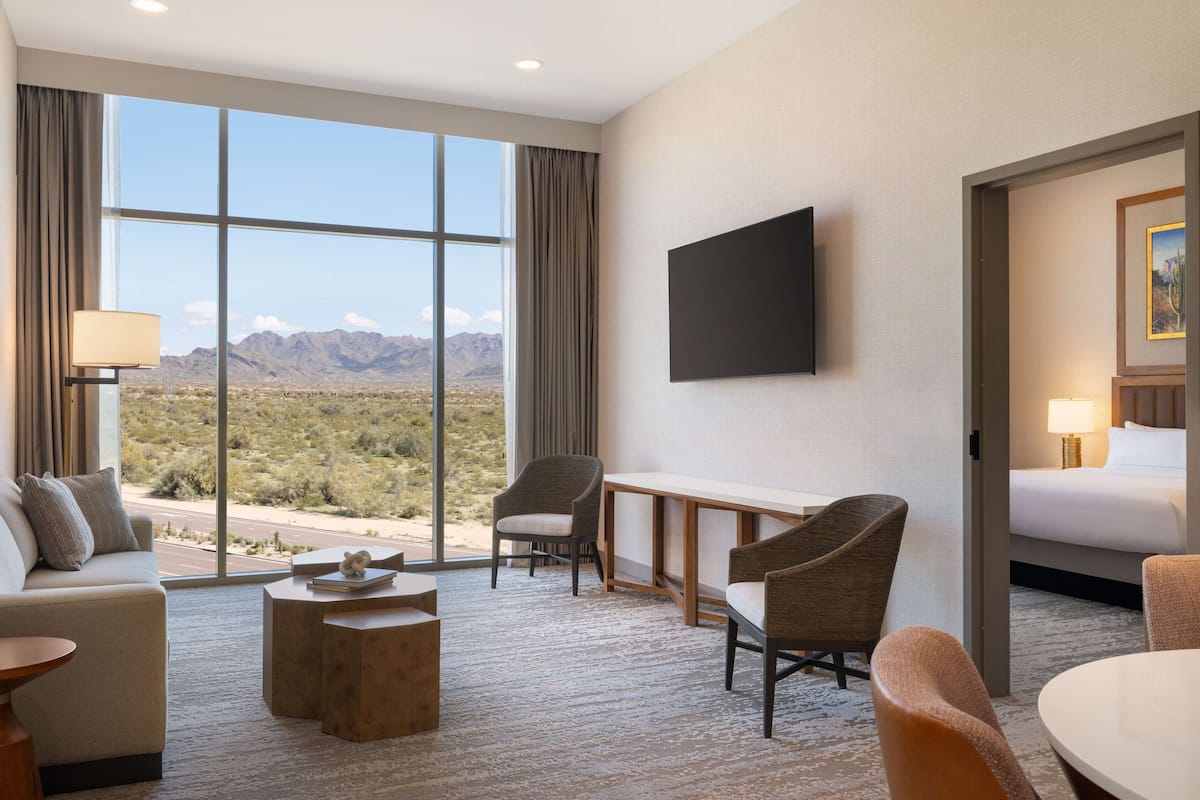 Ein Hotelzimmer mit großem Fenster und Blick auf die Wüste, einem wandmontierten Flachbildfernseher, einem Bett und einer Sitzecke mit Tisch und Stühlen.
