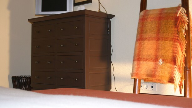 Una habitación con cama, cómoda con cajones, televisor encima y una lámpara.