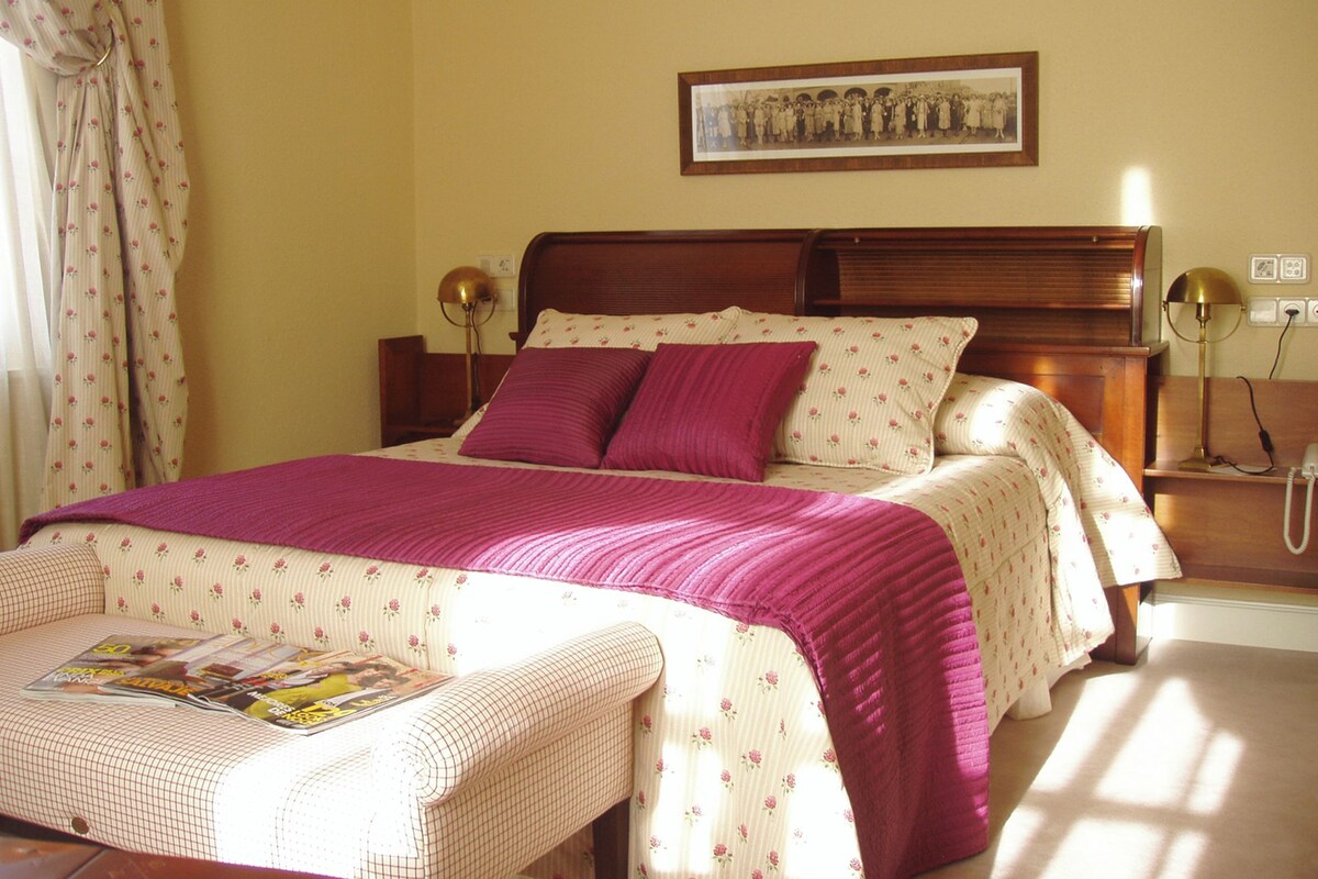 Un lit bien fait, avec une couverture et des oreillers roses, une table de chevet avec une lampe et un tableau encadré au mur.