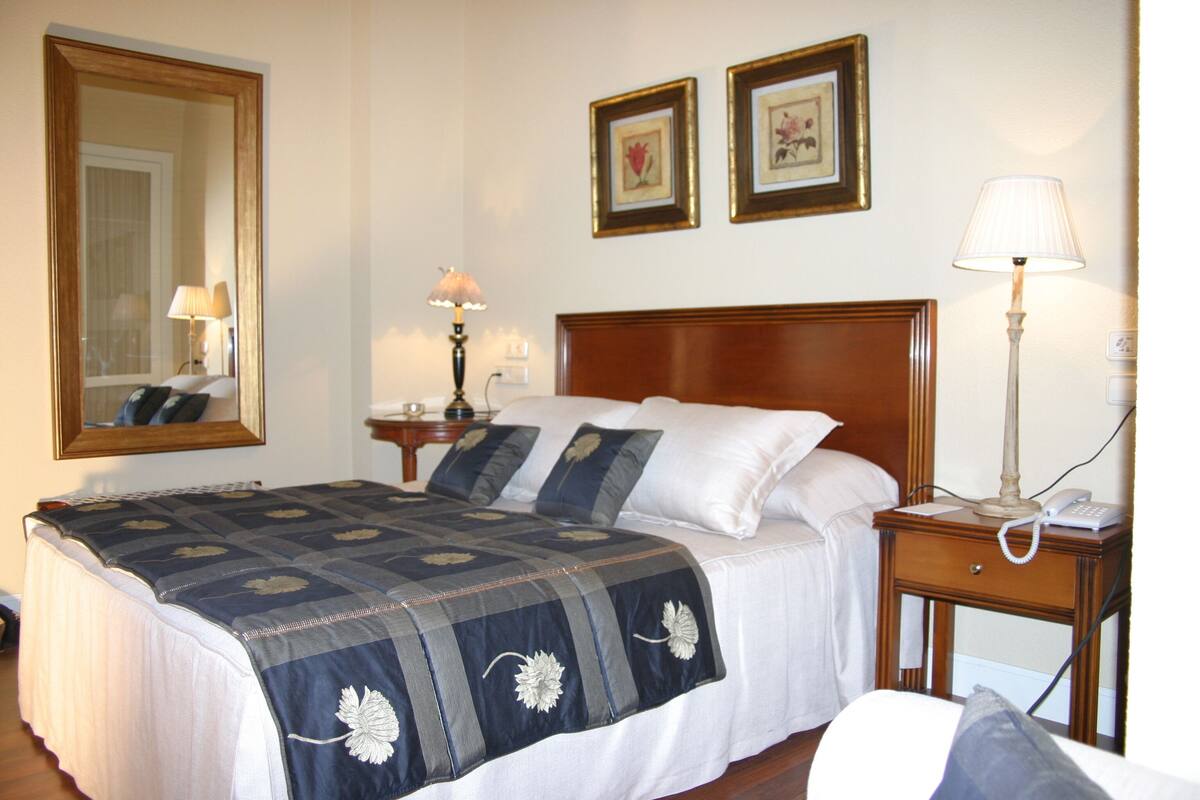 A neatly made bed with a blue and white quilt, a wooden headboard, and a bedside table with a lamp and a phone.