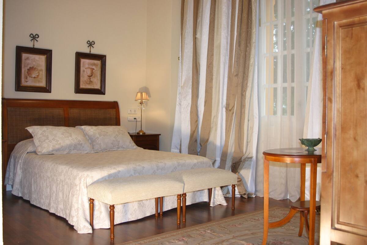 A bedroom with a large bed, a bedside table with a lamp, a wooden bench, and a small table with a bowl.
