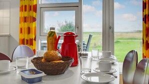 Ein Esstisch mit einem Brotkorb, einer Saftflasche und einer roten Karaffe, mit Blick auf ein grünes Feld durch ein Fenster.