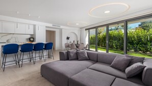 Una sala de estar moderna con un sofá seccional gris, una zona de comedor con una mesa pequeña y sillas, y una cocina con armarios blancos y taburetes de bar azules.