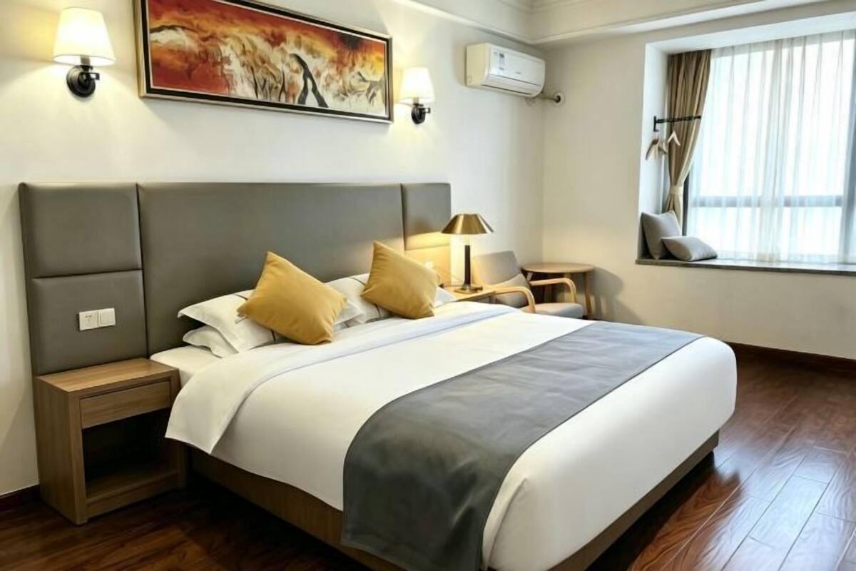 A hotel room with a large bed, bedside tables, a lamp, an air conditioning unit, and a window with curtains.