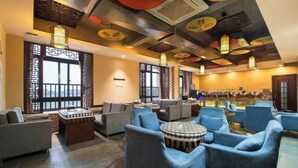 Un espace salon de style traditionnel, avec des canapés et des tables rondes, agrémenté d’un plafond orné d’éléments décoratifs et offrant une vue sur un coin cuisine à l’arrière-plan.
