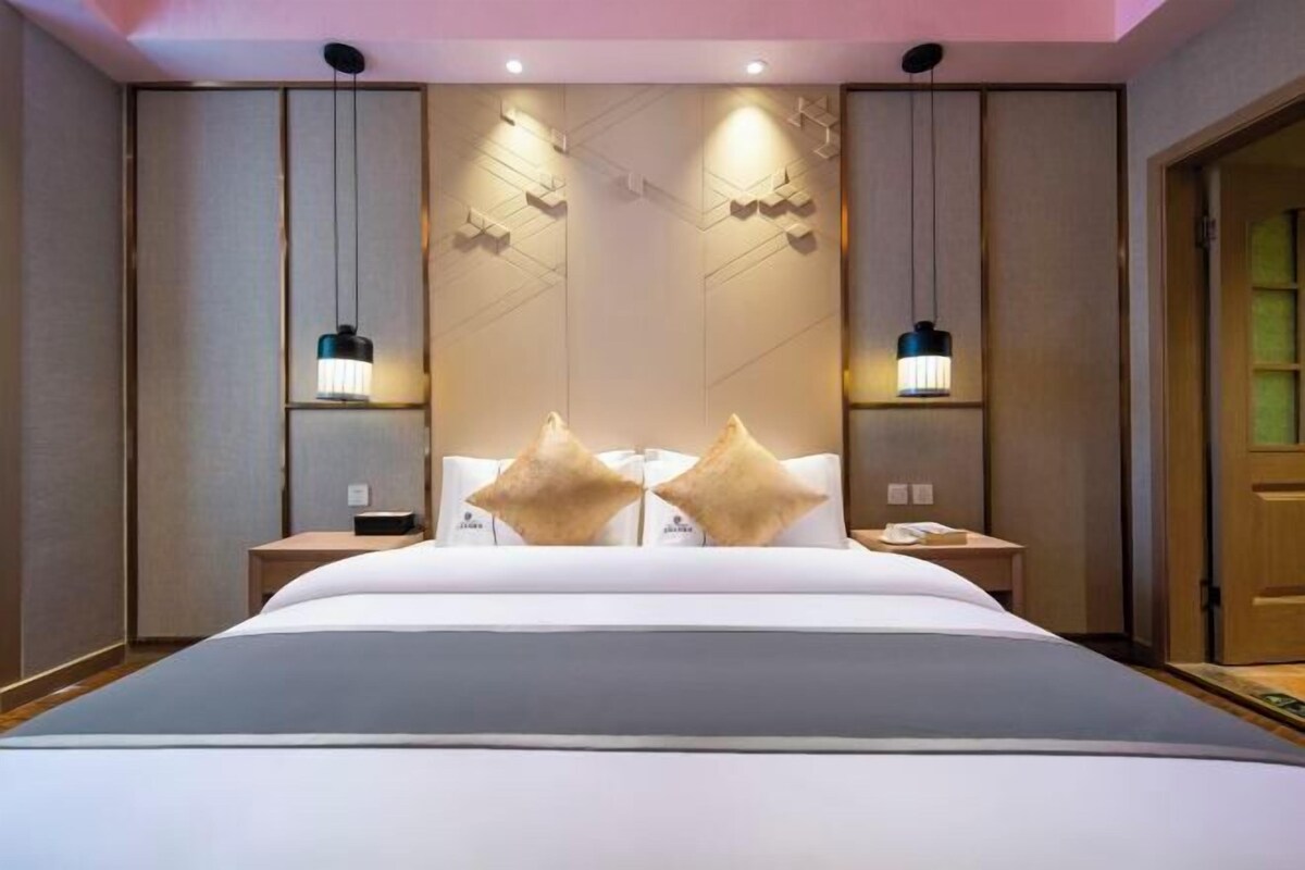 Habitación de hotel con una cama grande, dos mesitas de noche, una lámpara fijada a la pared y una puerta con un panel de vidrio verdoso.