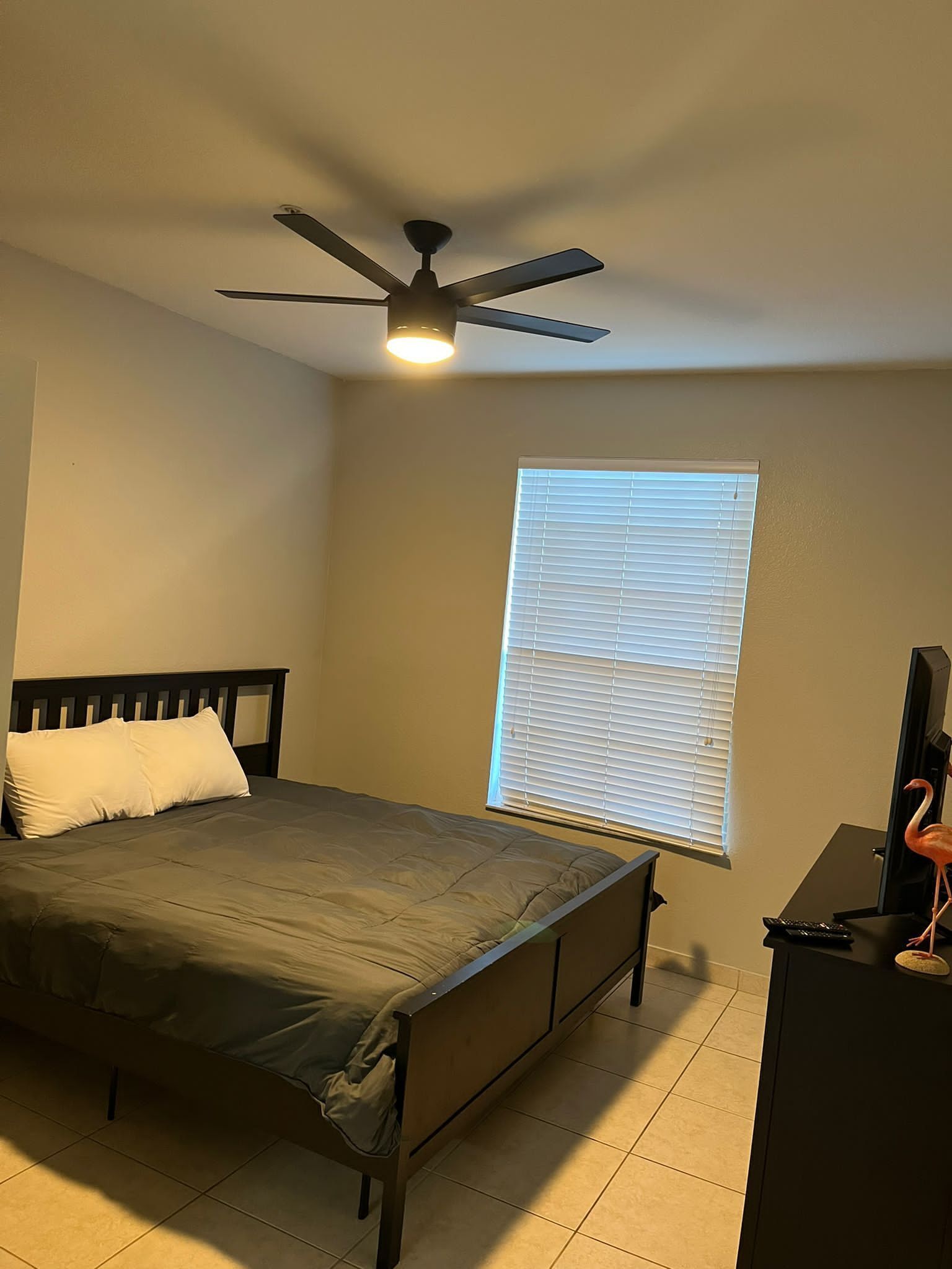 Une chambre à coucher comprenant un lit, un ventilateur de plafond, une fenêtre avec des stores et un meuble pour la télévision avec une télécommande.