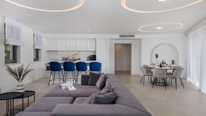 Un salon moderne avec un canapé gris, un coin repas avec une table ronde et des chaises, et une cuisine avec des meubles blancs et des chaises bleues.