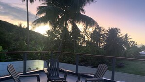 Ein Balkon mit vier Adirondack-Stühlen, einer Glasreling und Blick auf Palmen und einen Sonnenuntergang.