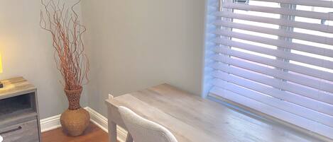 A room with a wooden desk, a chair, a vase with branches, a small table with a lamp, and a window with blinds.