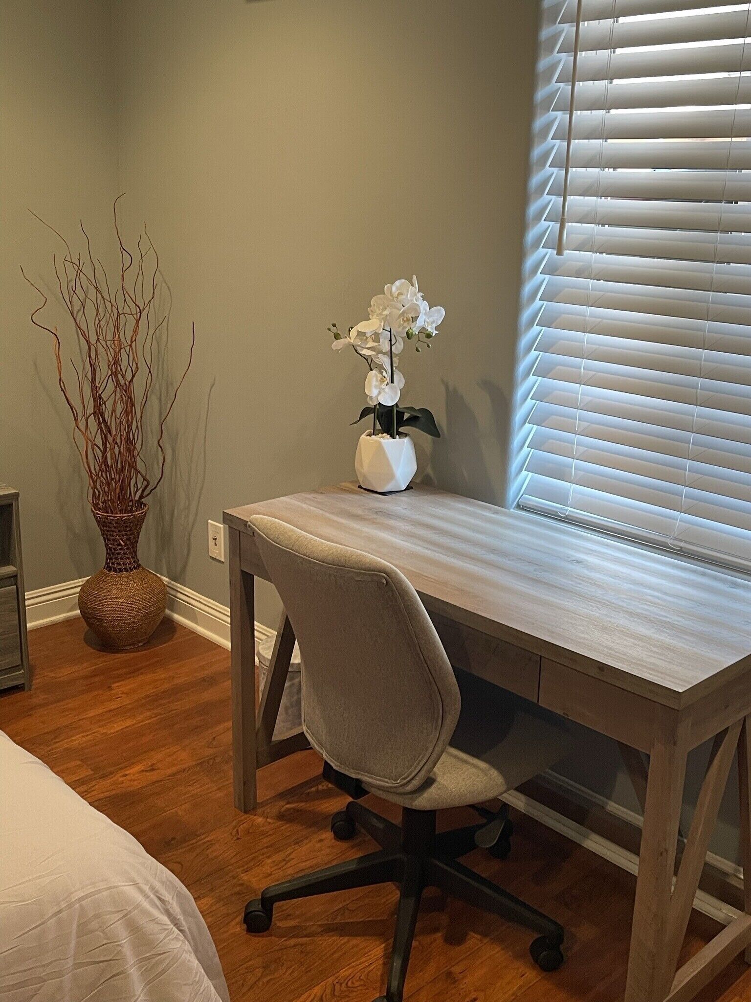 Un escritorio de madera con una silla, un jarrón blanco con una orquídea y una rama decorativa en un jarrón tejido.