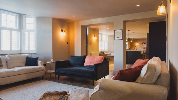 Ein Wohnzimmer mit einer Couch, Sesseln und einem Couchtisch. Im Hintergrund sind Fenster, eine Tür und ein Küchenbereich zu sehen.