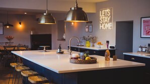 Eine moderne Küche mit einer großen Kochinsel, Barhocker, Pendelleuchten und einem Schild mit dem Aufdruck 'Let's Stay Home'.