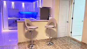 A modern kitchen with a gas stove, white cabinets, and blue under-cabinet lighting. There are two bar stools with cushioned seats. - RA-4 cómodo apartamento con vista al mar (Samaná)