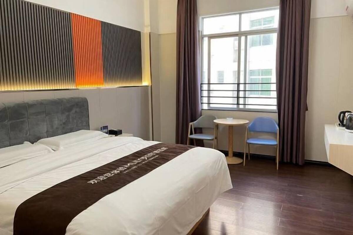 Une chambre d’hôtel avec un grand lit, une petite table, deux chaises, une fenêtre avec des rideaux et un panneau mural moderne.