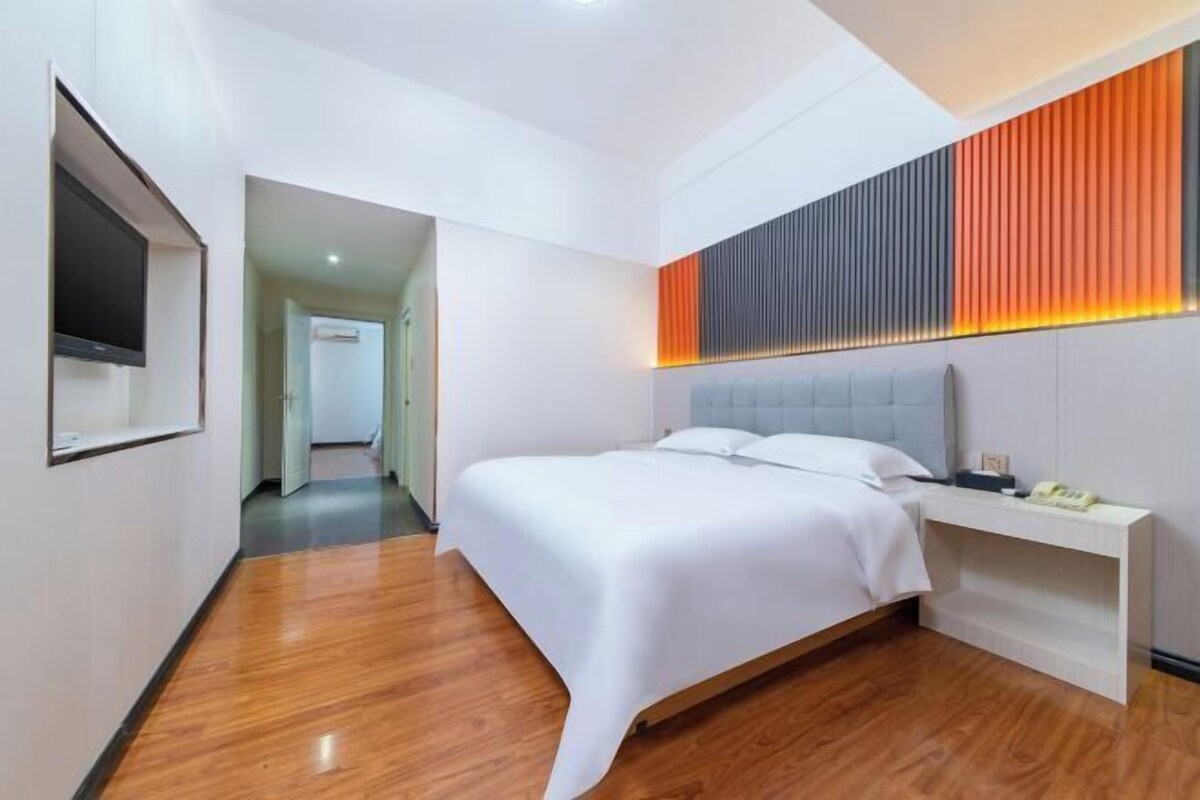 Une chambre d’hôtel moderne avec un grand lit, une table de chevet, un téléviseur fixé au mur et un couloir menant à une autre chambre.
