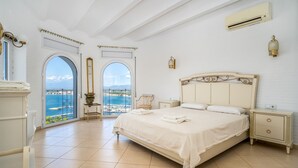Un dormitorio amplio con una cama grande, una mesita de noche y una silla. Se ve el mar a través de un ventanal de arco.