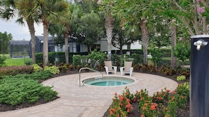 Una piscina circular con fuente de agua, rodeada por un patio empedrado, palmeras y plantas con flores.