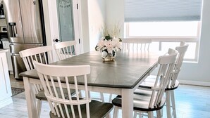 A dining area with a wooden table and white chairs, a kitchen in the background, and a window with blinds.