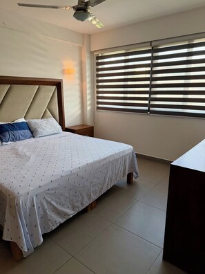 A bedroom with a bed, a nightstand, a desk, a ceiling fan, and a window with blinds.