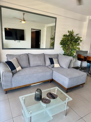 A modern living room with a sectional sofa, a large mirror, a flat-screen TV, a glass coffee table with decorative items, and a potted plant.