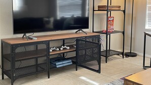 Una sala de estar moderna con un televisor de pantalla plana sobre un soporte de madera, una consola de videojuegos y un estante de metal negro con una planta y una caja de Jenga.