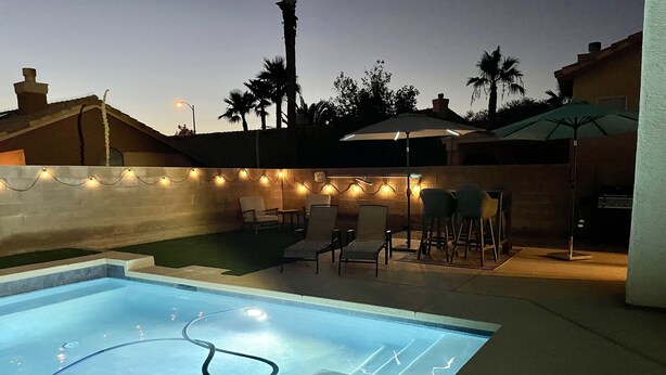 Una piscina con sillones de descanso y sombrillas, rodeada por un muro de ladrillo y palmeras.