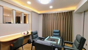 Quarto de hotel moderno com mesa de vidro, duas cadeiras de couro preto, uma secretária com cadeira, um espelho grande e uma janela coberta por cortinas.