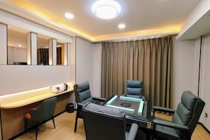 Quarto de hotel moderno com mesa de vidro, duas cadeiras de couro preto, uma secretária com cadeira, um espelho grande e uma janela coberta por cortinas.