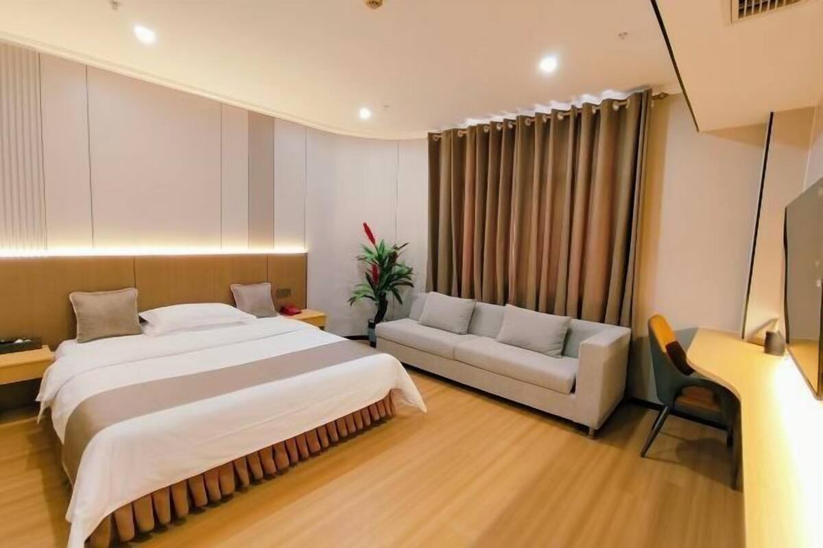 A modern hotel room with a large bed, a sofa, a desk, and a plant.