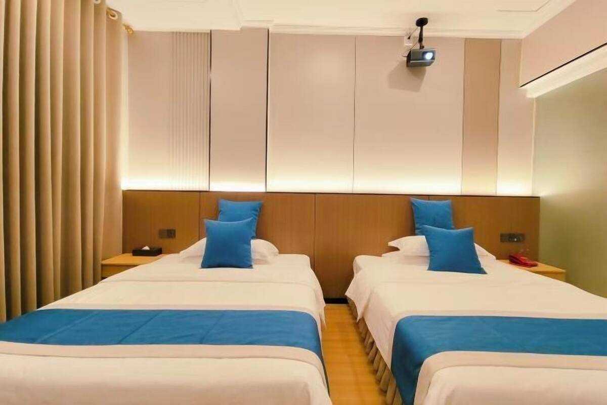A hotel room with two beds, a headboard with built-in lighting, a projector, and a curtain on the left.