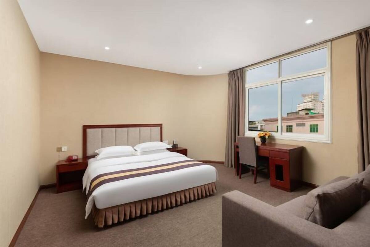 A hotel room with a bed, a desk, a chair, and a view of buildings through a window.