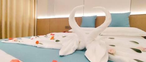 A hotel room with a bed covered in a blue sheet, white towels arranged in a heart shape, and scattered rose petals.