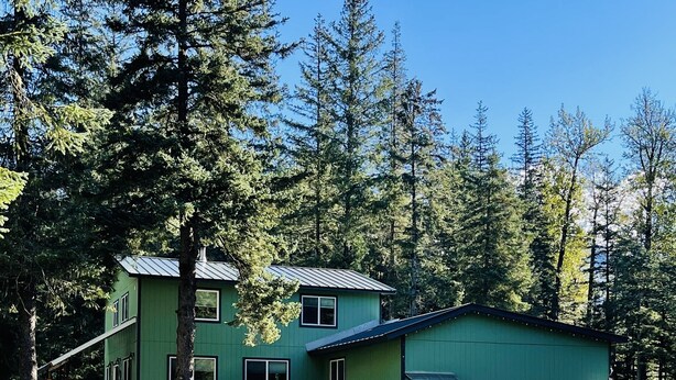 A green two-story house with a garage, surrounded by trees and a gravel driveway. - Private apartment in our home with separate entry. (Valdez)
