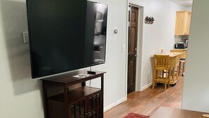 A living room with a flat-screen TV on a wooden stand, a dining area with a wooden table and chairs, and a kitchen in the background. - Private apartment in our home with separate entry. (Valdez)