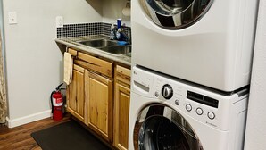 A compact kitchen with wooden cabinets, a double-door washer and dryer, a tiled backsplash, and a black rug. - Private apartment in our home with separate entry. (Valdez)