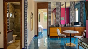 Una habitación de hotel moderna con baño, un comedor con una mesa redonda y dos sillas, una cocina con microondas y un espejo grande.