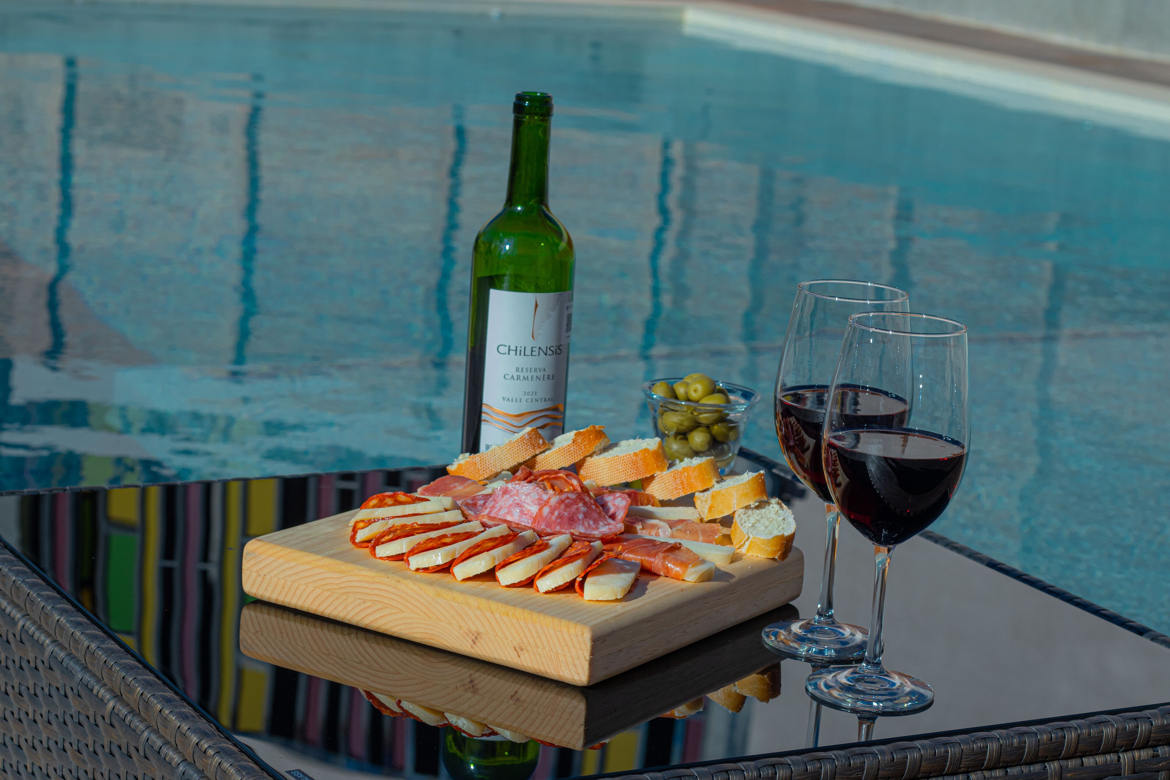 A glass table with a cheese board, wine glasses, and a bottle of wine by a pool.