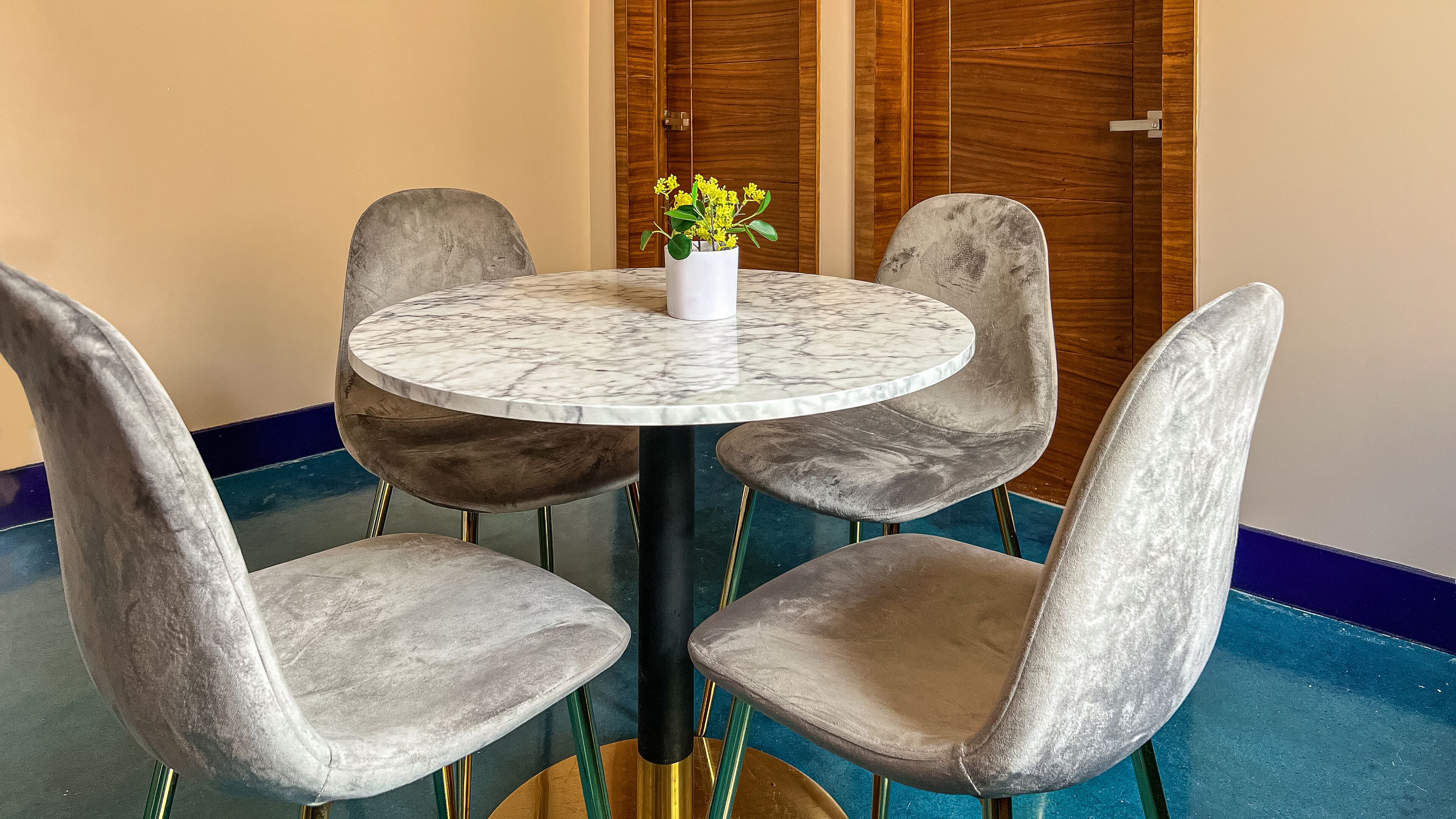 Une table ronde surmontée d’un plateau en marbre, entourée de quatre chaises grises à revêtement en tissu et à pieds métalliques. Un vase blanc contenant des fleurs jaunes est posé sur la table.