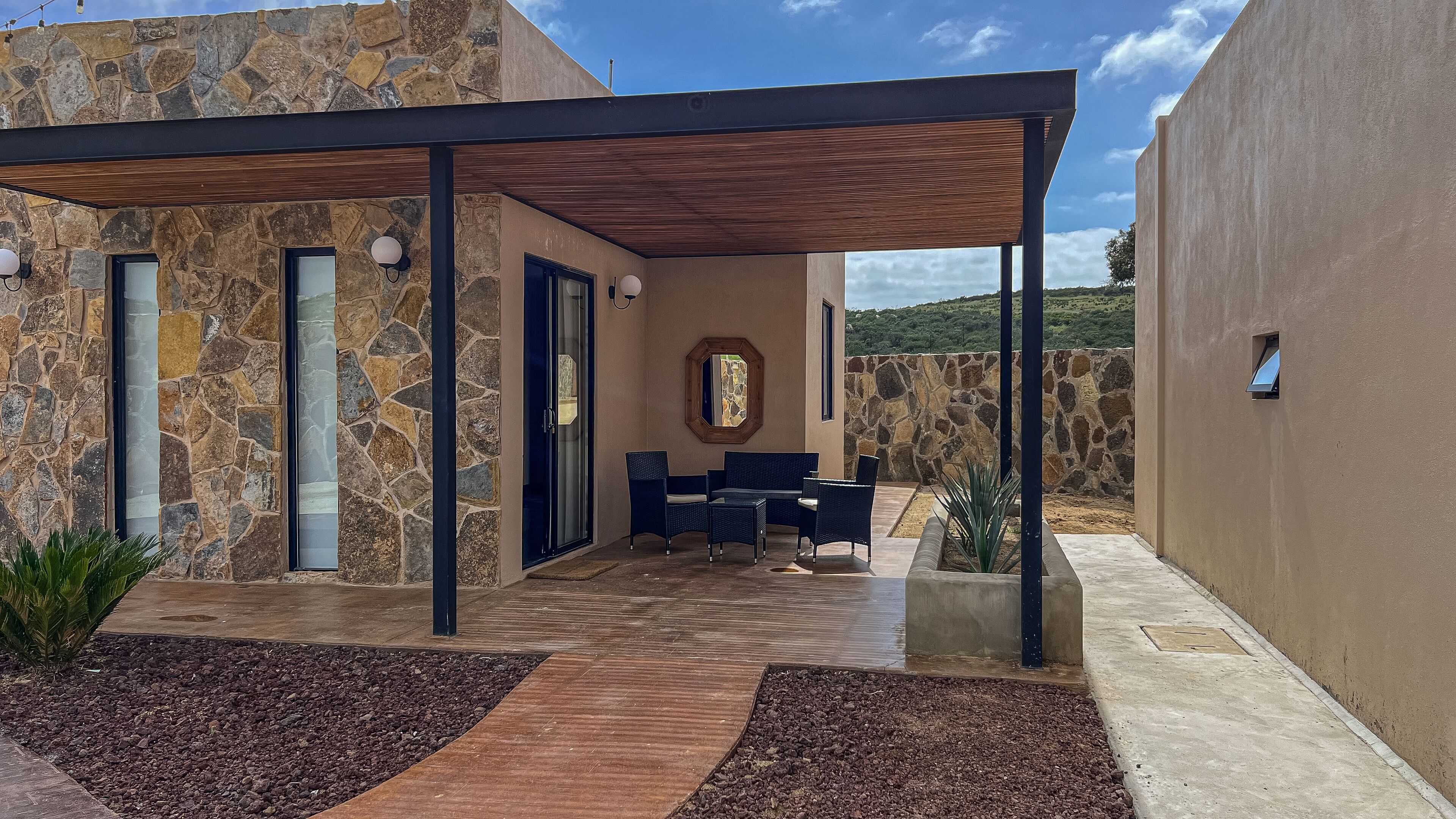 Un patio moderno al aire libre con paredes de piedra, una pérgola de madera y una zona de estar con sillas y una mesa.